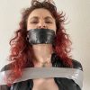 A very angry woman with red hair is gagged with black duct tape and tied up in a catsuit