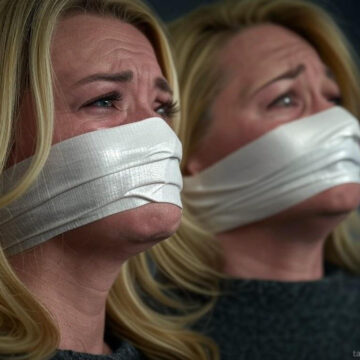 A blonde mother and her daughter sitting bound and gagged with white duct tape tightly wrapped around their mouths and noses, their eyes wide with fear and glistening as though on the verge of tears, illuminated by dim lighting in a tense and dramatic scene.