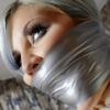 Blonde woman with silver and purple hair tightly gagged under shiny silver vinyl tape wrapped around her mouth