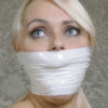 Blonde woman with blue eyes looking upward while tightly gagged under multiple layers of shiny white vinyl PVC tape