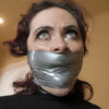 Brunette woman with curly dark hair looking upward while tightly gagged with shiny silver vinyl PVC tape wrapped around her mouth