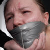 Crying woman with tears and snot running down her face, tightly gagged with shiny silver PVC tape while a hand holds her jaw