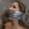 Woman in side profile with brown hair gagged under multiple layers of silver duct tape covering her mouth