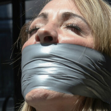 Blonde woman with eyes closed tightly gagged under multiple layers of shiny silver duct tape covering her mouth and chin