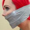 Red-haired woman in side profile tightly gagged with shiny silver PVC tape wrapped around her mouth