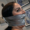 Dark-haired woman in side view gagged with multiple layers of silver duct tape covering her mouth and jaw