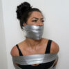 Brunette woman with hair in a high bun tightly gagged with wide silver duct tape covering her mouth, brows furrowed in discomfort