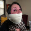 Brunette woman with purple eyeshadow tightly gagged with multiple layers of shiny white microfoam tape covering her mouth