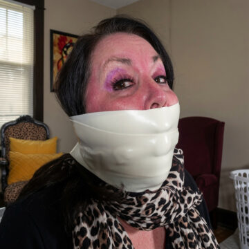 Brunette woman with purple eyeshadow tightly gagged with multiple layers of shiny white microfoam tape covering her mouth