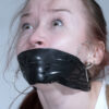 Young woman with freckles, septum piercing and large hoop earrings looking upward with a tear while tightly gagged with shiny black tape