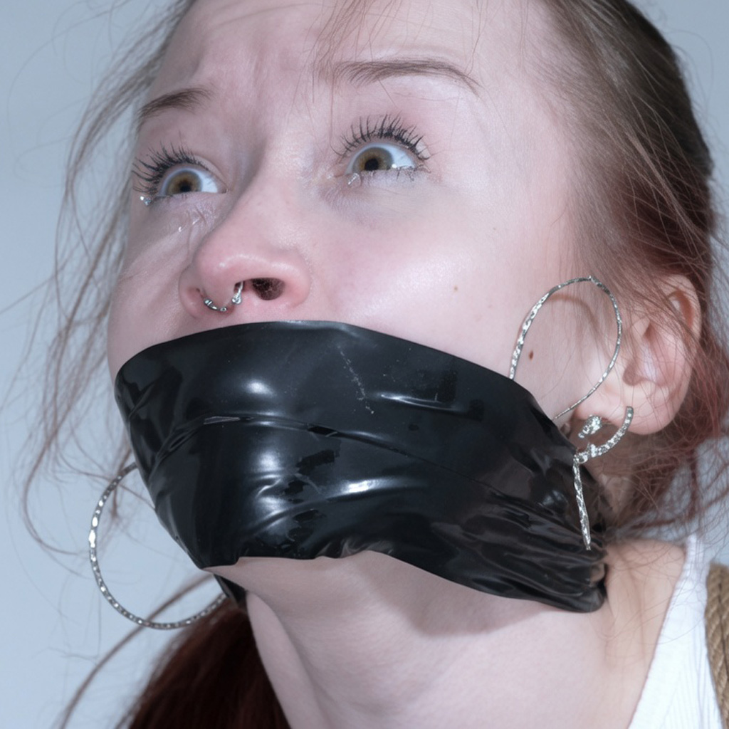 Young woman with freckles, septum piercing and large hoop earrings looking upward with a tear while tightly gagged with shiny black tape