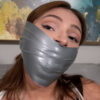 Young woman with reddish-brown hair and gold necklace gagged with wide silver duct tape covering her mouth in a tight wrap gag