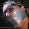 Woman wearing black-rimmed glasses with highlighted hair tightly gagged under shiny silver PVC tape