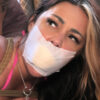 Woman with dark hair and blonde highlights looking upward while gagged with white medical tape over a sponge stuffing, rope around her chest